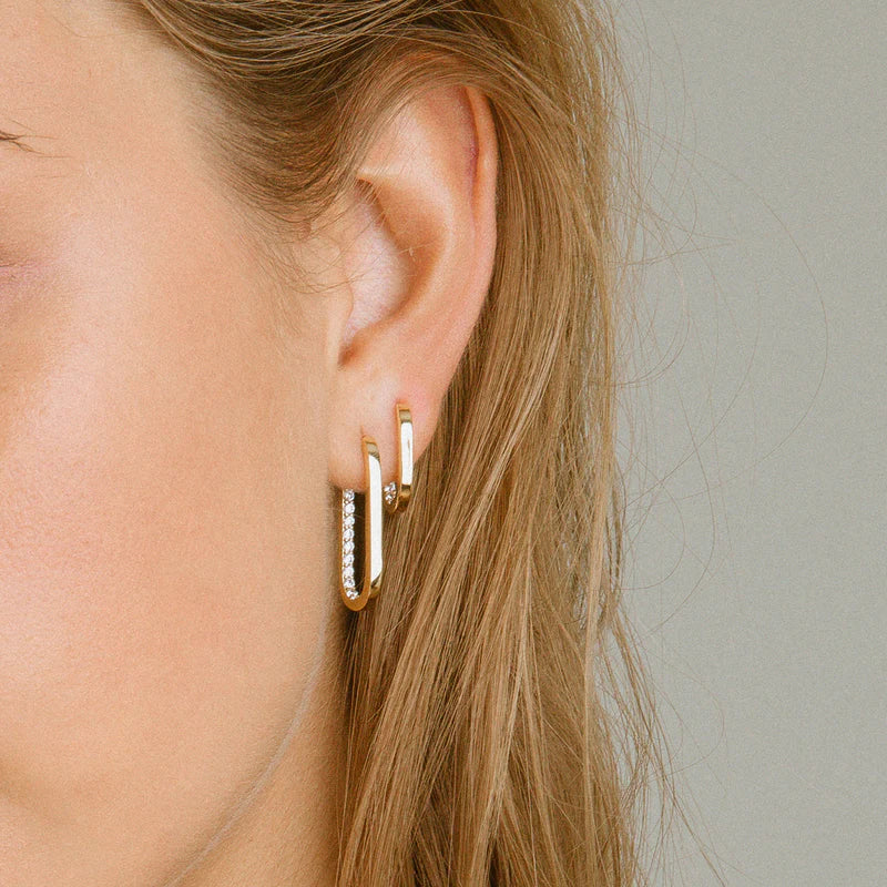 Close-up of a person wearing gold hoop earrings with clear stones against a neutral background